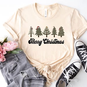 May include: A beige t-shirt with a graphic of five stylized Christmas trees with ornaments and a star on top. The text "Merry Christmas" is printed in black below the trees.