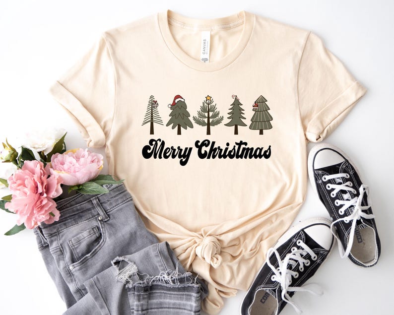 May include: A beige t-shirt with a graphic of five stylized Christmas trees with ornaments and a star on top. The text "Merry Christmas" is printed in black below the trees.