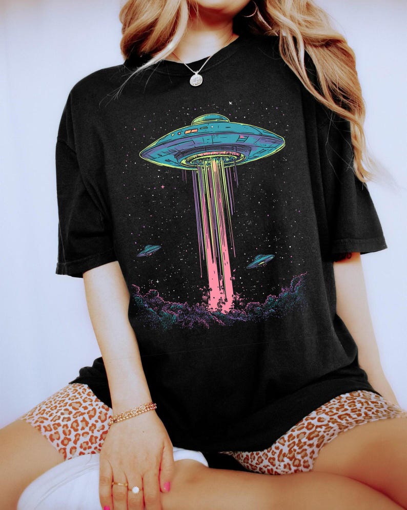 May include: Black t-shirt with a vibrant graphic of a UFO beaming light onto a starry background. Two additional UFOs are depicted. The design is set against a backdrop of dark clouds.
