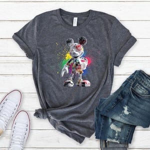 May include: A grey t-shirt with a colourful graphic of Mickey Mouse in a graffiti style. The design features vibrant paint splatters and a unique artistic representation of the iconic character. The shirt is paired with blue jeans and white trainers.