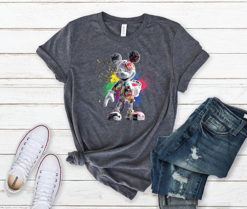 May include: A grey t-shirt with a colourful graphic of Mickey Mouse in a graffiti style. The design features vibrant paint splatters and a unique artistic representation of the iconic character. The shirt is paired with blue jeans and white trainers.