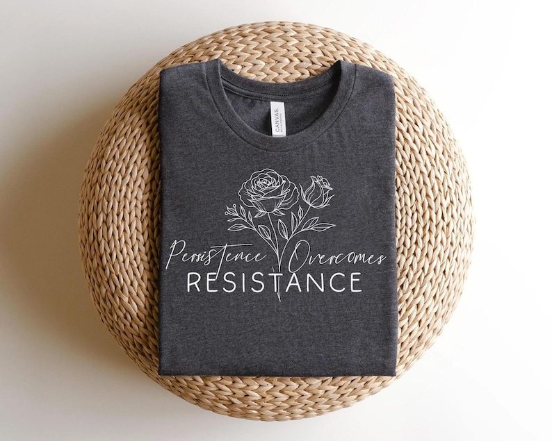 May include: Dark gray t-shirt with a white floral design and the words "Persistence Overcomes Resistance". The shirt is folded and placed on a woven, circular surface.