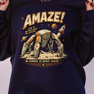 May include: A dark navy blue sweatshirt featuring a graphic print of a stone-textured robotic crab on a lunar surface. The design includes a small rocket, planets, stars, and the text AMAZE! LIFE IS BETTER WHEN YOU EXPLORE and BE CURIOUS. BE BRAVE. AMAZE!