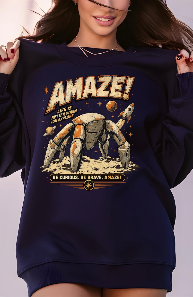 May include: A dark navy blue sweatshirt featuring a graphic print of a stone-textured robotic crab on a lunar surface. The design includes a small rocket, planets, stars, and the text AMAZE! LIFE IS BETTER WHEN YOU EXPLORE and BE CURIOUS. BE BRAVE. AMAZE!