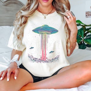 May include: A cream-coloured t-shirt with a vibrant UFO design. The graphic depicts a flying saucer with a beam of pink and purple light, and two smaller UFOs. This casual t-shirt is suitable for everyday wear and features a unique design.