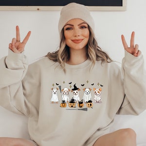 Op de afbeelding: Een beige sweatshirt met een afbeelding van vijf honden die verkleed zijn als geesten voor Halloween. De honden houden pompoenen en een bezem vast. Het sweatshirt heeft een ronde hals en lange mouwen.