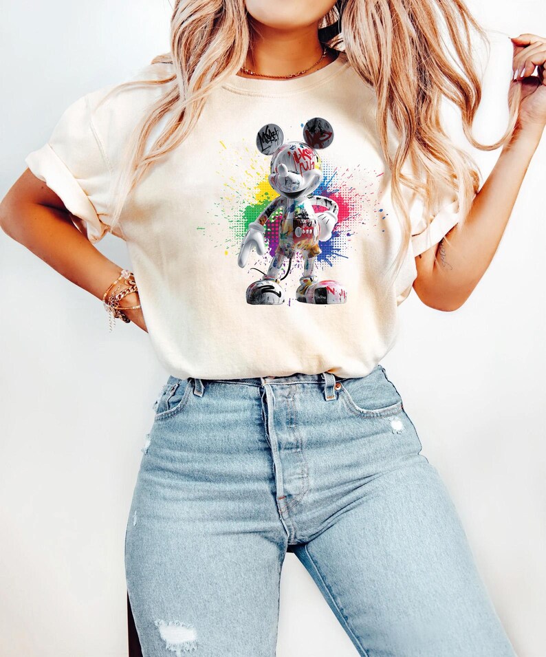 May include: A cream-coloured t-shirt with a colourful graphic of Mickey Mouse in a graffiti style. The design includes a vibrant splash of paint in various colours. The shirt is worn with light-wash blue jeans.