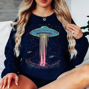May include: Navy blue sweatshirt with a vibrant graphic of a UFO beaming light downwards. The design includes a starry night sky and two smaller UFOs. A comfortable, casual style.
