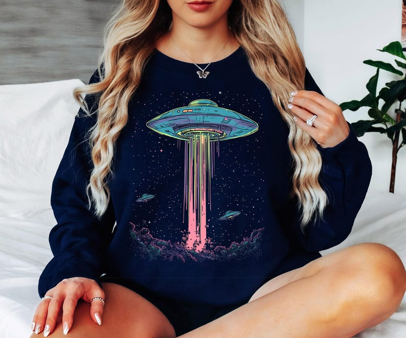 May include: Navy blue sweatshirt with a vibrant graphic of a UFO beaming light downwards. The design includes a starry night sky and two smaller UFOs. A comfortable, casual style.