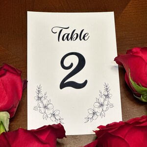 Op de afbeelding: Witte kaart met zwarte tekst die "Tafel 2" luidt met een bloemmotief aan de onderkant. De kaart is omringd door rode rozen.