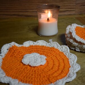 May include: A lit white candle in a glass holder sits near a stack of handmade crochet coasters. The coasters have an orange center with a white scalloped edge. One coaster is in the foreground.