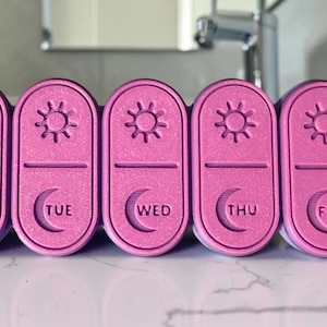 May include: A set of seven pink pill organizers arranged in a row, each featuring a sun icon at the top and a moon icon above the day of the week abbreviation. The labels include SUN, MON, TUE, WED, THU, FRI, and SAT, with a textured finish.
