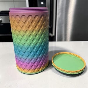 May include: A cylindrical container with a textured, diamond-patterned exterior featuring a rainbow gradient of purple, pink, orange, yellow, green, and blue. The container has a matching circular lid with a green top and an orange threaded rim.