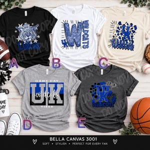 May include: Five short-sleeved crew neck t-shirts featuring various blue and white graphic designs related to the Kentucky Wildcats. The shirts are displayed in black, white, cream, heather gray, and dark charcoal gray colors with text.