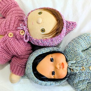 Könnte beinhalten: Zwei Puppen mit gehäkelten Outfits. Die obere Puppe hat helle Haut, braunes Haar sowie eine rosa gehäkelte Haube und einen Pullover mit Holzknöpfen. Die untere Puppe hat schwarzes Haar sowie eine hellblaue gehäkelte Haube und einen Pullover.