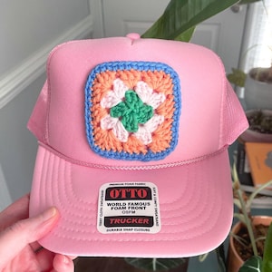May include: A pink trucker hat with a crocheted square patch. The patch features a floral design with green, white, orange, and blue colors. The hat has a foam front and a snap closure. The hat is labeled "OTTO" and "TRUCKER".