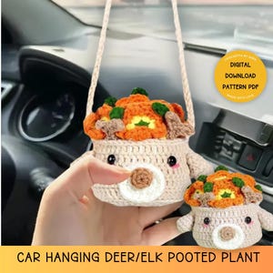 May include: Crocheted car hanging ornament featuring a deer or elk with a pot of orange and green flowers on its head. The ornament is made of beige yarn and has a cute face. The text "CROCHETTA BY SARA DIGITAL DOWNLOAD PATTERN PDF MADE WITH LOVE" is visible in the image. The text "CAR HANGING DEER/ELK POOTED PLANT" is also visible in the image.