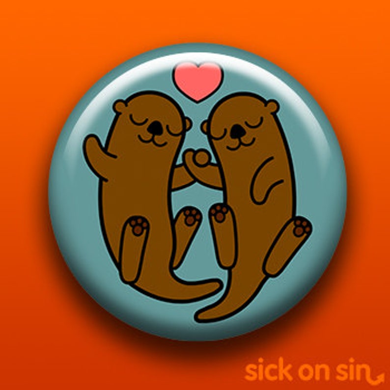 K&ouml;nnte beinhalten: Ein runder Button mit t&uuml;rkisblauem Hintergrund, der zwei Cartoon-Otter zeigt, die sich unter einem rosa Herzen an den H&auml;nden halten. Die Otter sind braun mit schwarzen Details. Der Button hat eine gl&auml;nzende Oberfl&auml;che und einen Schatten.