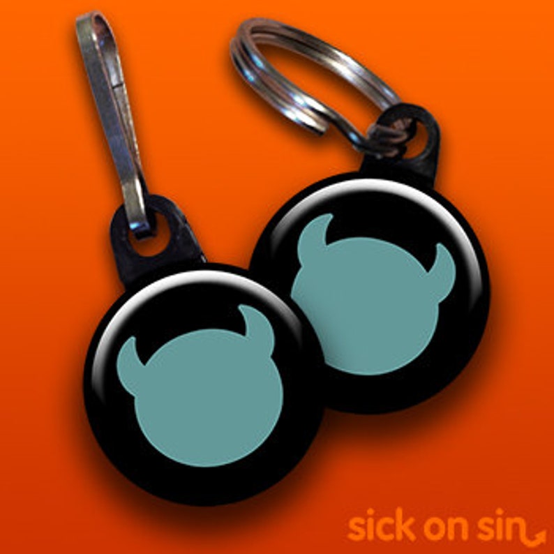 Op de afbeelding: Twee zwarte sleutelhangers met een zilveren metalen ring. Elke sleutelhanger heeft een ronde, zwarte knop met een lichtblauw silhouet van een cartoonmonster met hoorns.