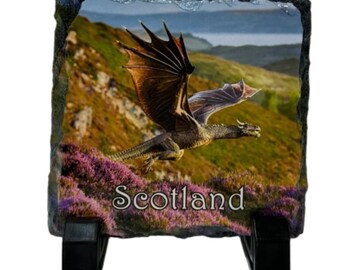 Puede incluir: Una baldosa decorativa de pizarra con un dragón volando sobre un paisaje escocés, con brezo morado y colinas. La palabra "Scotland" está impresa en blanco. La baldosa está sostenida por un soporte negro.
