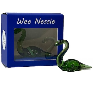 Puede incluir: Una figura de Nessie de vidrio verde junto a una caja azul con el texto "Wee Nessie". La figura es una representación estilizada del monstruo del lago Ness. La caja tiene una ventana transparente.