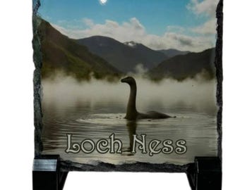 Puede incluir: Placa rectangular de pizarra con una imagen del lago Ness, mostrando al monstruo emergiendo del agua. El paisaje incluye montañas y un cielo nublado, con la inscripción "Loch Ness" en la parte inferior. Soporte negro.