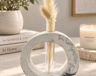 May include: A circular white concrete vase with a textured, marbled black finish at the base. A clear glass test tube is inserted into the center of the ring, holding two beige, feathery dried botanical stems that extend upward from the opening.