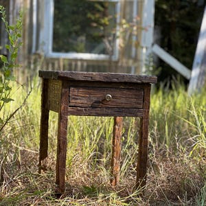 May include: A rustic wooden end table with a single drawer and a metal knob. The table is made of reclaimed wood and has a natural finish. It is sitting in a grassy area.