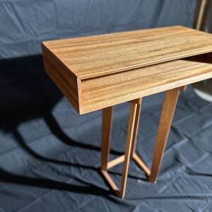 Peut inclure: Une table d'appoint en bois faite à la main, avec une finition brun rougeâtre chaleureuse. La table présente un plateau rectangulaire avec une étagère inférieure et des pieds inclinés, mettant en valeur un design moderne. Ses dimensions sont d'environ 61 cm de long, 30 cm de large et 71 cm de haut.