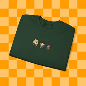 May include: A dark green t-shirt with a pixelated design of three characters. The characters are wearing brown, black, and green clothing.