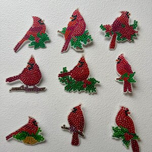 Puede incluir: Una colección de nueve parches decorativos con pájaros cardenales rojos adornados con pequeñas piedras de estrás brillantes. Cada ave se muestra en varias posturas, algunas posadas sobre hojas o ramas verdes, exhibiendo un plumaje rojo vibrante.