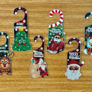 Könnte beinhalten: Eine Sammlung von Türhängern zum Thema Weihnachten mit verschiedenen Designs. Jeder Hänger zeigt eine festliche Figur oder ein Element, darunter ein Rentier, ein Weihnachtsbaum, der Weihnachtsmann und ein Schneemann. Die Hänger tragen die Texte "Merry Christmas" und "Merry & Bright".