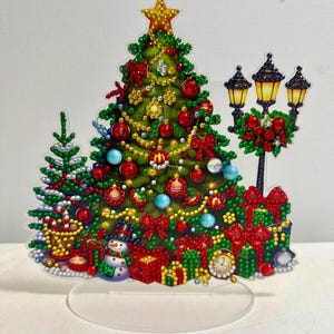May include: A festive Christmas tree decoration with a star on top, adorned with red and blue ornaments, bows, and lights. Presents, a snowman, and a street lamp are also featured. The decoration is mounted on a clear, oval base.