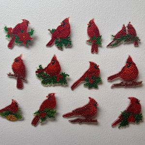 Puede incluir: Una colección de doce adornos individuales de pájaros cardenales elaborados con cuentas facetadas rojas y verdes. Cada pieza presenta una pose distinta de un cardenal rojo posado sobre follaje o ramas verdes, creando un aspecto texturizado.