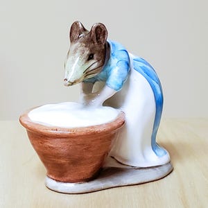 May include: A ceramic figurine of a mouse wearing a blue and white outfit, leaning over a brown bowl filled with a white substance. The mouse has brown ears and a light-colored face. The figurine is on a gray base.