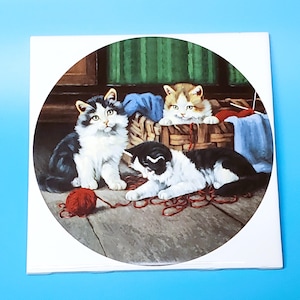 May include: A round decorative plate featuring three kittens playing with red yarn. One kitten sits, two others are in a basket. The kittens have black, white, and orange fur. The background includes a basket, blue fabric, and green curtains.