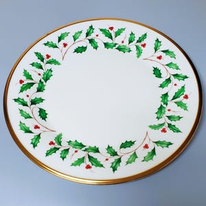 May include: White ceramic plate with a gold rim, decorated with a green holly and red berry wreath design. The plate is round and has a smooth surface, perfect for serving holiday meals. The holly wreath is painted around the edge of the plate.