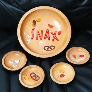 May include: A large wooden serving bowl and four smaller matching wooden bowls. The large bowl features the red text SNAX with illustrations of popcorn, pretzels, and fruit slices. The smaller bowls feature individual illustrations of snacks.