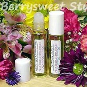 May include: Two clear glass roller bottles with white caps and labels. The bottles are filled with a light yellow liquid. The bottles are surrounded by a variety of colorful flowers.