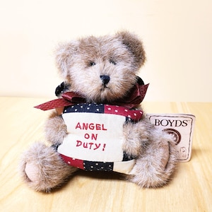 Peut inclure: Un ours en peluche en fourrure brune, portant un n&oelig;ud bordeaux et tenant une banni&egrave;re patriotique avec l'inscription "ANGEL ON DUTY!". Une &eacute;tiquette "BOYDS" est visible. L'ours est assis sur une surface en bois.