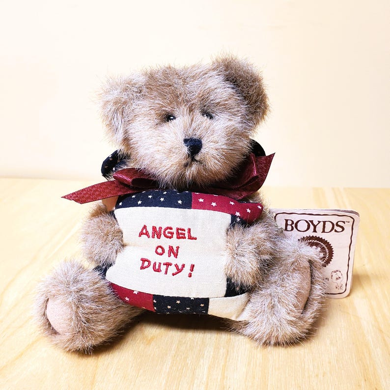 Peut inclure: Un ours en peluche en fourrure brune, portant un n&oelig;ud bordeaux et tenant une banni&egrave;re patriotique avec l'inscription "ANGEL ON DUTY!". Une &eacute;tiquette "BOYDS" est visible. L'ours est assis sur une surface en bois.