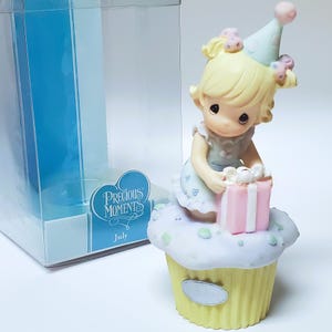 May include: A Precious Moments figurine of a girl holding a pink gift box atop a cupcake. The figurine has blonde hair and wears a party hat. The cupcake is yellow with white frosting and green sprinkles. The box reads "July".