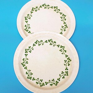 May include: Two round ceramic plates with a speckled off-white finish. Each plate features a circular pattern of green shamrock vines printed along the inner rim. The plates are arranged vertically, one slightly overlapping the other on a blue background.