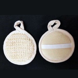 May include: A white round bath sponge with a textured surface on one side and a smooth surface on the other side. The sponge has a white elastic band around the smooth side.