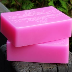 May include: Two stacked, bright pink rectangular soaps. The top soap has a floral design etched into its surface. The soaps are smooth and have a glossy appearance. The background is blurred, with green foliage and a dark wooden surface.