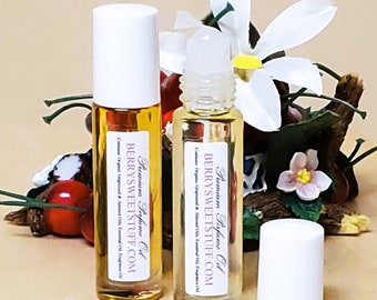 May include: Two clear glass roller bottles with white caps and labels. The labels read "Premium Perfume Oil" and "BerrySweetStuff.com". The bottles are sitting on a wooden surface with a floral arrangement in the background.