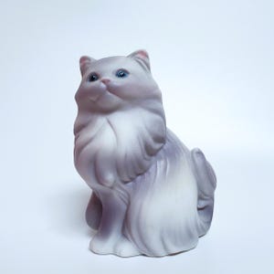 May include: A ceramic cat figurine featuring long, flowing fur in shades of soft white and muted lavender-gray. The cat is depicted in a seated position with its head tilted upward, showcasing bright blue eyes and a detailed, sculpted facial expression.