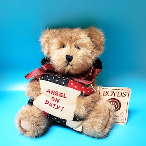 Peut inclure: Un ours en peluche brun avec un n&oelig;ud rouge et une banni&egrave;re avec l'inscription "ANGEL ON DUTY!". La banni&egrave;re pr&eacute;sente un motif d'&eacute;toiles et de rayures. L'ours porte une &eacute;tiquette "BOYDS".