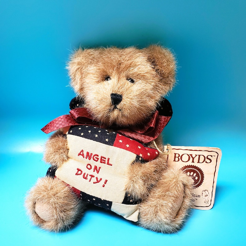 Peut inclure: Un ours en peluche brun avec un n&oelig;ud rouge et une banni&egrave;re avec l'inscription "ANGEL ON DUTY!". La banni&egrave;re pr&eacute;sente un motif d'&eacute;toiles et de rayures. L'ours porte une &eacute;tiquette "BOYDS".