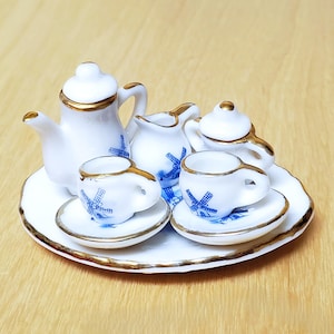 May include: A miniature white porcelain tea set arranged on a matching round tray. The set includes a teapot, a creamer, a sugar bowl, and two cups with saucers. Each piece features a blue windmill pattern and delicate gold trim accents.